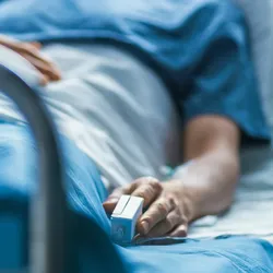 Photo of a clinical trial participant lying in a hospital bed covered with a sheet and wearing an oximeter on their index finger.