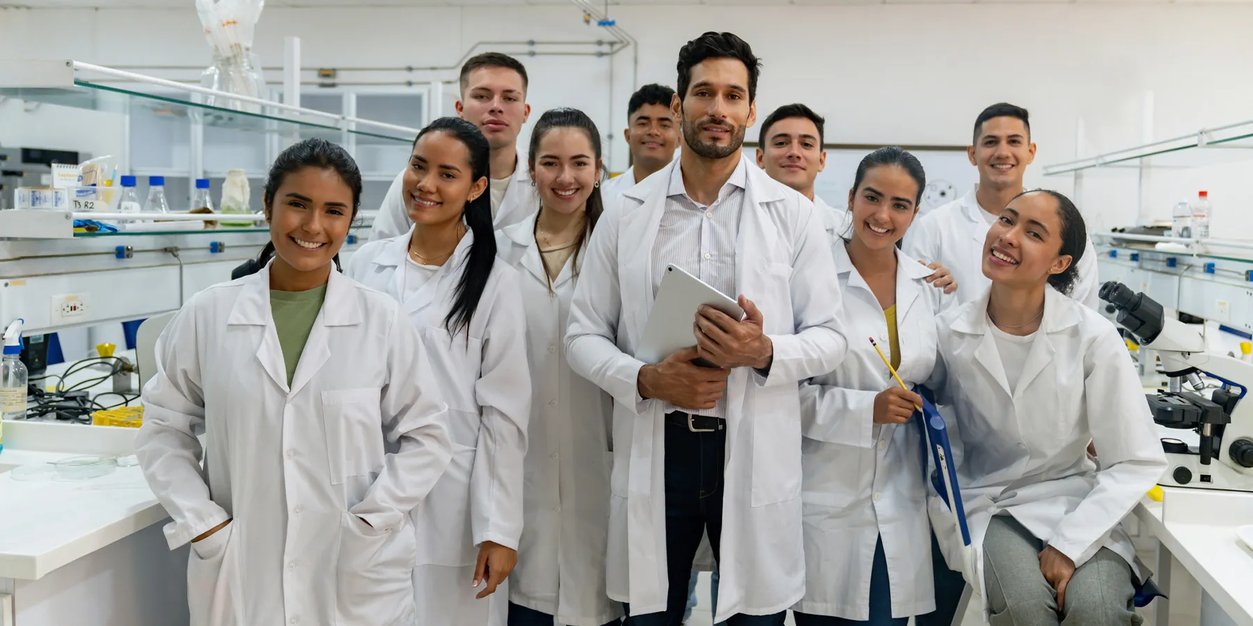 Photo of a group scientists wearing white lab coats standing in a laboratory and smiling at the camera.