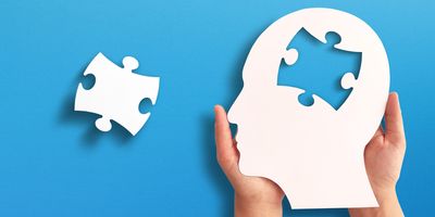 Photo of two hands holding a white cut-out of a human head profile with a puzzle piece missing from the brain on a blue background.