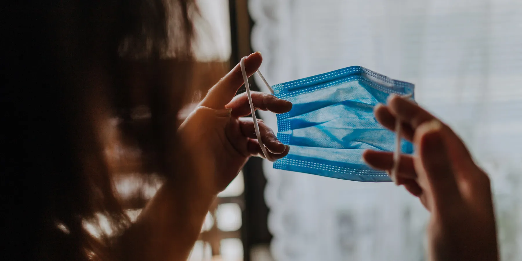 Photo of a woman holding a blue surgical mask up to her face.