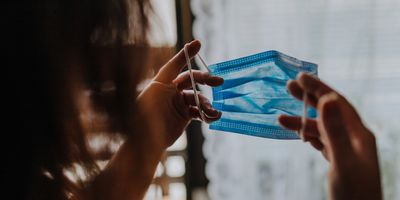 Photo of a woman holding a blue surgical mask up to her face.