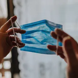 Photo of a woman holding a blue surgical mask up to her face.