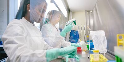 Photo of two young female medical laboratory scientists wearing PPE and working at a biological safety cabinet in a laboratory setting, with concern on their face for the recent science and healthcare policy changes in the US.