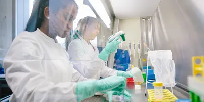Photo of two young female medical laboratory scientists wearing PPE and working at a biological safety cabinet in a laboratory setting, with concern on their face for the recent science and healthcare policy changes in the US.