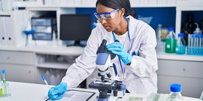 A photo of a young Black woman who is a medical laboratory scientist wearing a white lab coat using a microscope in the clinical laboratory.