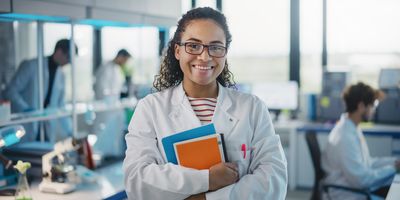  Photo of a Black woman clinical laboratory professional smiling and wearing a white lab coat while holding a clipboard during a health and safety and ergonomics meeting.