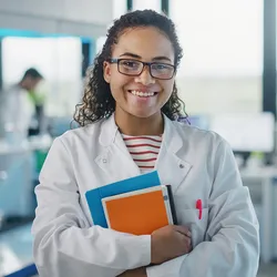  Photo of a Black woman clinical laboratory professional smiling and wearing a white lab coat while holding a clipboard during a health and safety and ergonomics meeting.