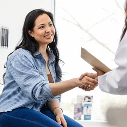 Photo of a young female cancer patient with long black hair in a health care setting shaking the hand of a healthcare provider conducting cancer screening.