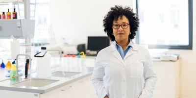 Photo of a medical laboratory professional wearing a white lab coat and standing in the clinical lab with their hands in their pockets.