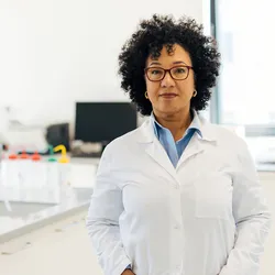 Photo of a medical laboratory professional wearing a white lab coat and standing in the clinical lab with their hands in their pockets.