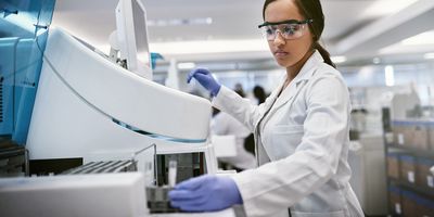 Photo of a clinical lab professional wearing a white lab coat and gloves in a clinical lab and loading a multiple sclerosis sample into an analyzer.