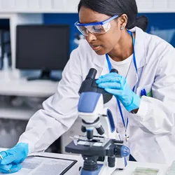 Photo of a young Black woman scientist wearing PPE and using a microscope to examine breast cancer tissues in a clinical laboratory.