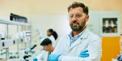 Photo of a male clinical laboratory professional wearing a white lab coat and blue gloves standing with his arms crossed in a medical laboratory. 