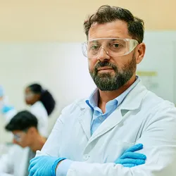 Photo of a male clinical laboratory professional wearing a white lab coat and blue gloves standing with his arms crossed in a medical laboratory. 