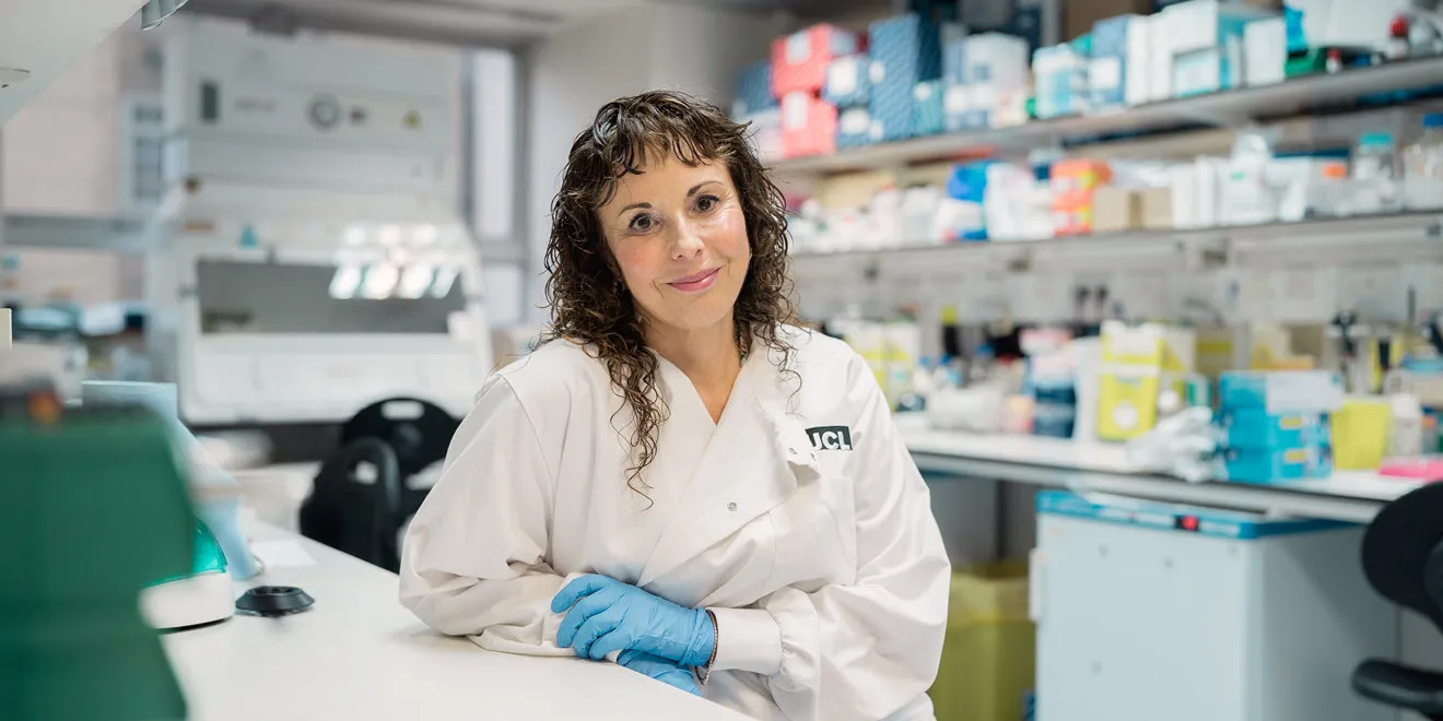 Photo of Sarah Tabrizi wearing a white laboratory coat and blue gloves while sitting at a laboratory bench.