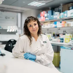 Photo of Sarah Tabrizi wearing a white laboratory coat and blue gloves while sitting at a laboratory bench.