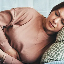 Woman experiencing abdominal discomfort on a couch due to gaps in follow-up testing.